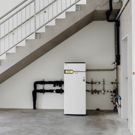 Ein weißer Wasserboiler steht in einem Raum mit einer Treppe im Hintergrund.