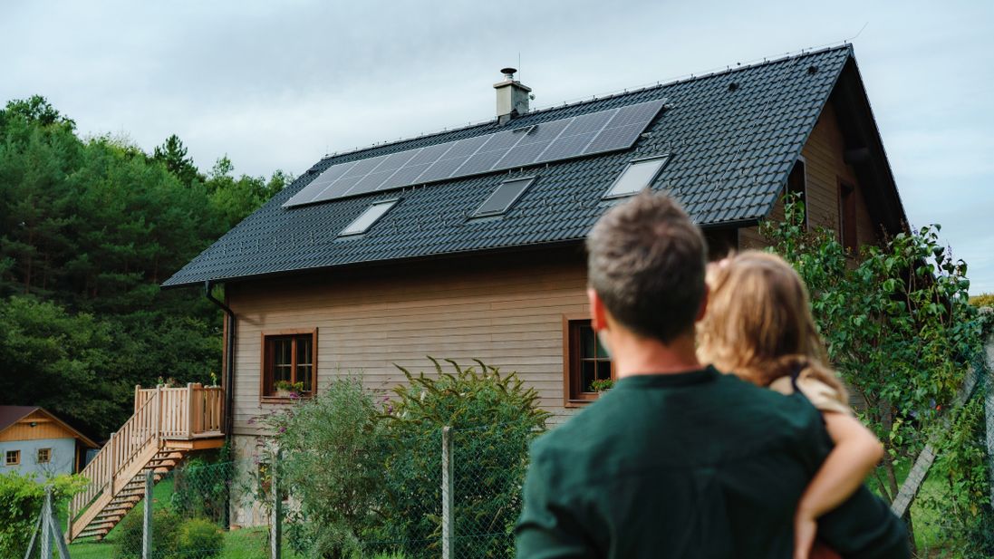 Familienvater mit Tochter auf dem Arm schaut auf ein Haus mit Photovoltaik-Anlagen auf dem Dach